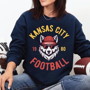May include: Navy blue sweatshirt with a graphic featuring a wolf wearing sunglasses and a red beanie. The text reads "Kansas City Football 1980" in yellow and red. A football is visible.