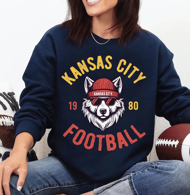 May include: Navy blue sweatshirt with a graphic featuring a wolf wearing sunglasses and a red beanie. The text reads "Kansas City Football 1980" in yellow and red. A football is visible.