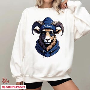 May include: White sweatshirt featuring a cartoon ram wearing a blue beanie with "Los Angeles" text, sunglasses, and a blue hoodie. The ram has large curved horns and a brown and tan face. The sweatshirt is a casual style.
