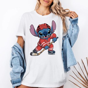 May include: White t-shirt featuring a cartoon Stitch character in a red hockey uniform, helmet, and skates, holding a hockey stick. The character is blue with large ears and a mischievous grin. The shirt is a casual, everyday wear item.