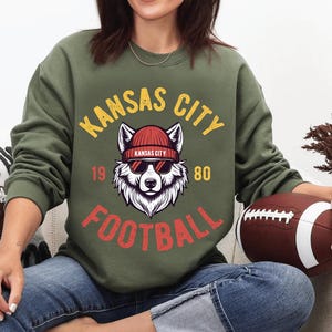 May include: Olive green sweatshirt with a graphic featuring a wolf wearing sunglasses and a red beanie. The text reads "KANSAS CITY FOOTBALL 1980" in yellow and red. A brown football is held in the person's hand.