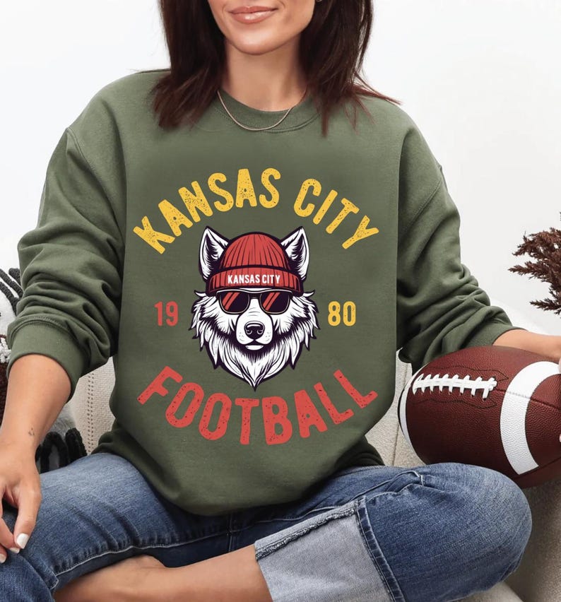 May include: Olive green sweatshirt with a graphic featuring a wolf wearing sunglasses and a red beanie. The text reads "KANSAS CITY FOOTBALL 1980" in yellow and red. A brown football is held in the person's hand.