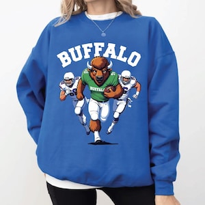 May include: Royal blue sweatshirt with the word "BUFFALO" in white block letters. The graphic features a buffalo mascot running with a football, flanked by two football players in white uniforms. The mascot wears a green jersey.