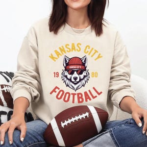 May include: Cream-colored sweatshirt with a graphic featuring a wolf wearing sunglasses and a beanie, with the text "Kansas City Football 1980". A brown football with white stripes is in the foreground.