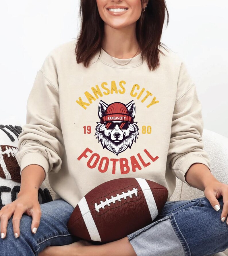 May include: Cream-colored sweatshirt with a graphic featuring a wolf wearing sunglasses and a beanie, with the text "Kansas City Football 1980". A brown football with white stripes is in the foreground.