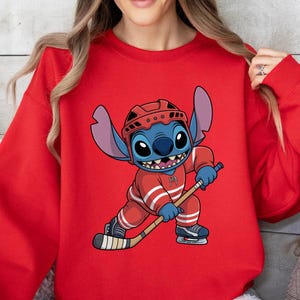 May include: Red sweatshirt featuring a cartoon character in a hockey uniform. The character is blue with large ears and is holding a hockey stick. The hockey uniform is red with white stripes. The character is wearing a red helmet and ice skates.