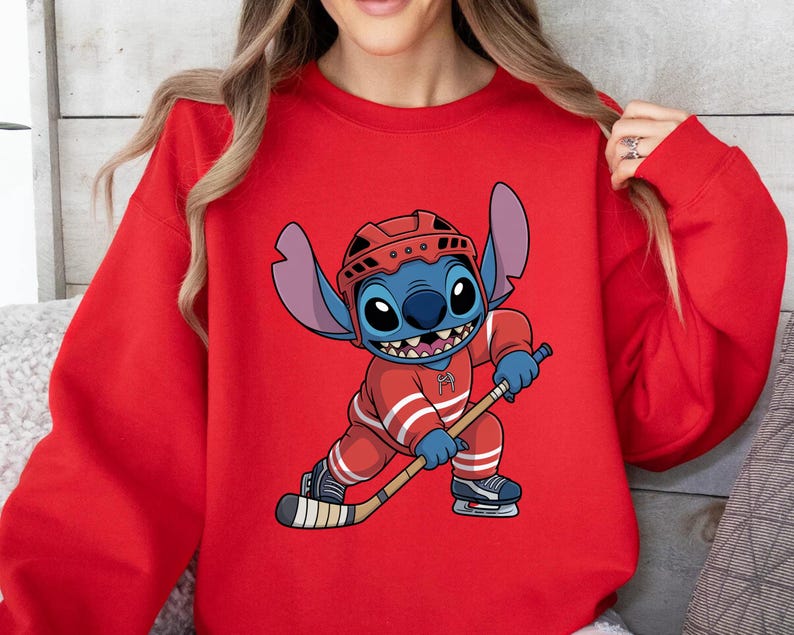 May include: Red sweatshirt featuring a cartoon character in a hockey uniform. The character is blue with large ears and is holding a hockey stick. The hockey uniform is red with white stripes. The character is wearing a red helmet and ice skates.