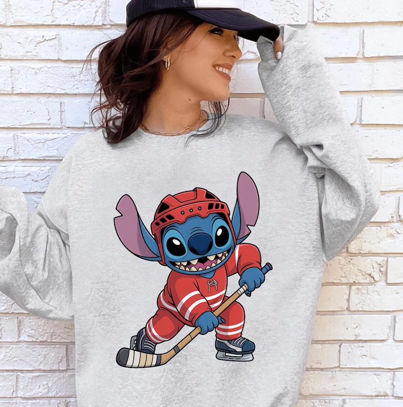 May include: A light gray sweatshirt featuring a cartoon Stitch character in a red hockey uniform and helmet, holding a hockey stick. The character is blue with large ears and a wide grin. The sweatshirt has a crew neck.