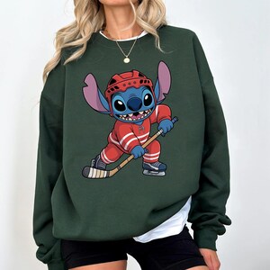 May include: Dark green sweatshirt featuring a cartoon character in a red hockey uniform, helmet, and skates, holding a hockey stick. The character is blue with large ears and a wide grin. The sweatshirt has a white collar.