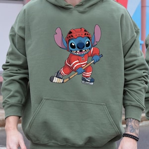 May include: Olive green hoodie featuring a cartoon character in a red hockey uniform with white stripes, a red helmet, and ice skates. The character is holding a hockey stick and appears to be skating. The hoodie has a kangaroo pocket.
