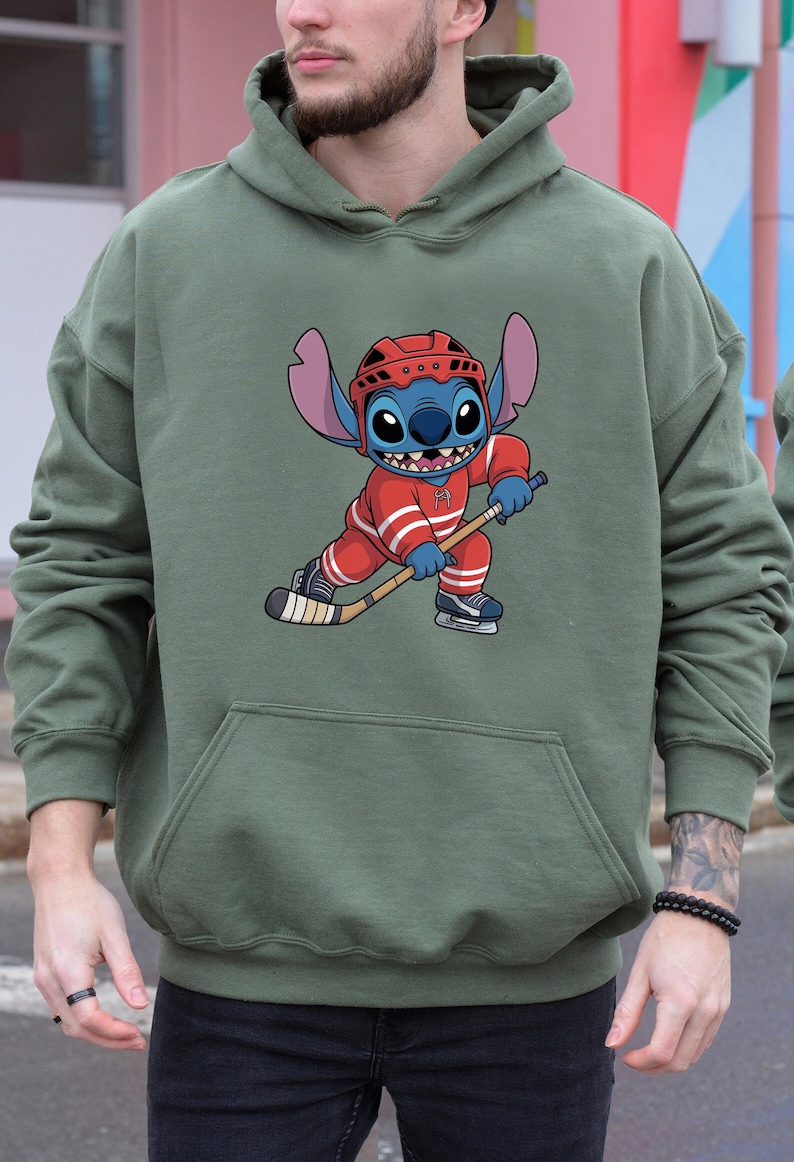 May include: Olive green hoodie featuring a cartoon character in a red hockey uniform with white stripes, a red helmet, and ice skates. The character is holding a hockey stick and appears to be skating. The hoodie has a kangaroo pocket.