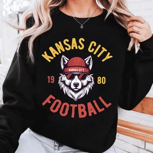 May include: Black sweatshirt with a graphic featuring a wolf wearing sunglasses and a beanie. The text "KANSAS CITY FOOTBALL" is arched above and below the wolf illustration, with "1980" on either side.