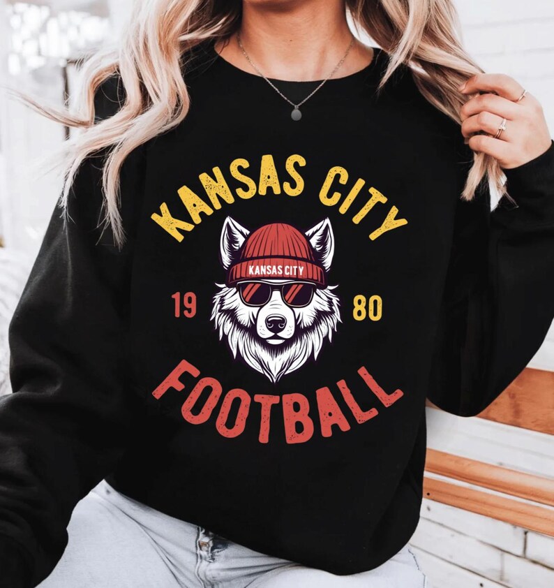 May include: Black sweatshirt with a graphic featuring a wolf wearing sunglasses and a beanie. The text "KANSAS CITY FOOTBALL" is arched above and below the wolf illustration, with "1980" on either side.