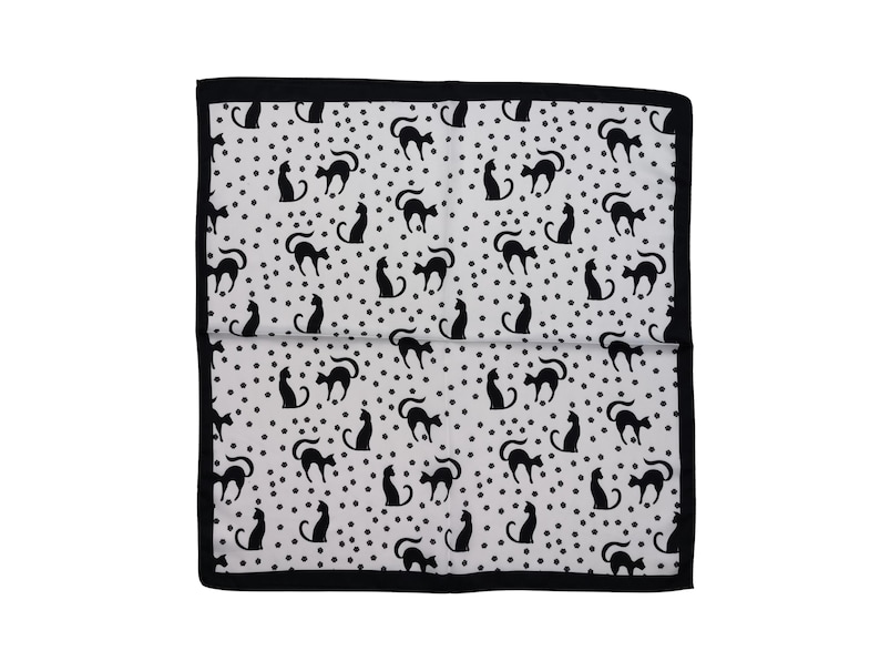 May include: A white and black patterned textile featuring a repeating design of black cat silhouettes and small black dots. The edges are bordered with a solid black trim. The textile is folded in half.