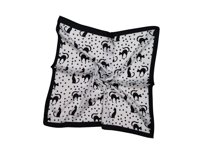 May include: Square white scarf with a black border, featuring a pattern of black cat silhouettes and small black polka dots. The scarf is folded and twisted, showcasing the design.