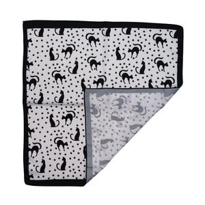 May include: A white and black patterned textile featuring black cat silhouettes and small black dots. The textile has a black border and a folded corner revealing a similar pattern in gray and white.