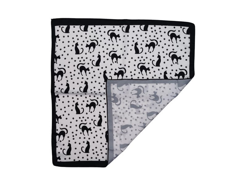 May include: A white and black patterned textile featuring black cat silhouettes and small black dots. The textile has a black border and a folded corner revealing a similar pattern in gray and white.