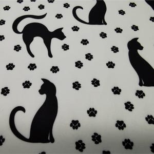 May include: White fabric with a repeating pattern of black cat silhouettes and paw prints. The cats are in various poses, some sitting and others standing. The paw prints are scattered across the fabric.