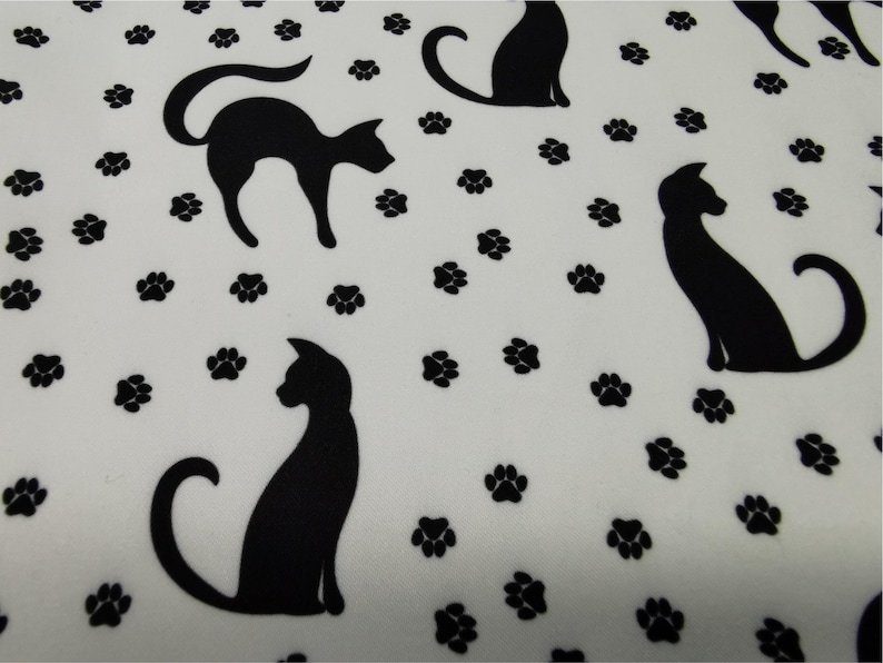 May include: White fabric with a repeating pattern of black cat silhouettes and paw prints. The cats are in various poses, some sitting and others standing. The paw prints are scattered across the fabric.