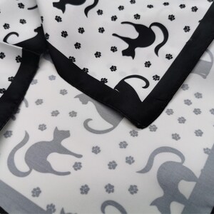 May include: White fabric with a black border, featuring a repeating pattern of black cat silhouettes and gray paw prints. The fabric is folded, showing the pattern and border details.