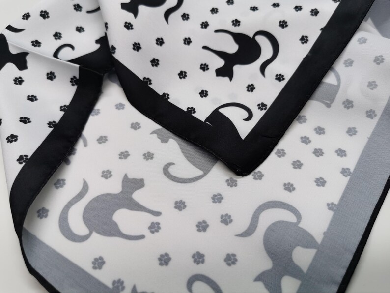 May include: White fabric with a black border, featuring a repeating pattern of black cat silhouettes and gray paw prints. The fabric is folded, showing the pattern and border details.