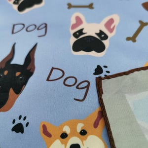 May include: Light blue fabric with repeating dog-themed illustrations. Features various dog breeds, paw prints, and the word "Dog" in brown. A portion of a light blue and brown-edged square is visible.