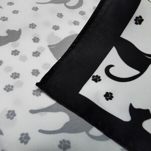 May include: A white fabric with a repeating pattern of gray cat silhouettes and black paw prints. A corner of the fabric is folded over, featuring a black border and a black cat silhouette.