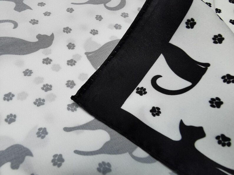 May include: A white fabric with a repeating pattern of gray cat silhouettes and black paw prints. A corner of the fabric is folded over, featuring a black border and a black cat silhouette.