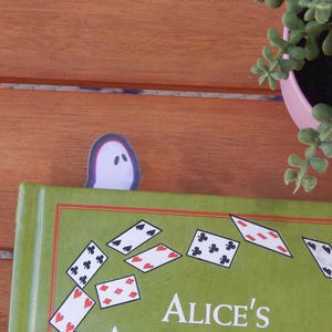 May include: A green book titled "Alice's Adventures in Wonderland" with a white ghost-shaped bookmark. The book has playing card illustrations. A pink pot with a green plant is in the background.