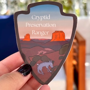 May include: A sticker with a landscape design and the words "Cryptid Preservation Ranger." The sticker features a desert scene with red rock formations, a blue sky, and a light purple creature. The sticker is shaped like a shield.