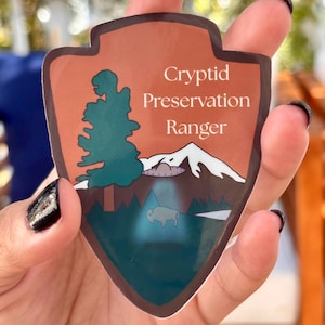 May include: A shield-shaped sticker with a brown border and background. The sticker features a green tree, white mountains, a gray UFO, and a bison. The text "Cryptid Preservation Ranger" is in white. The sticker is held in a hand.