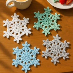 May include: Four handmade crochet snowflake coasters in white, light blue, gray, and mint green are displayed on a wooden surface. A white mug with a heart-shaped latte art and a plate of pancakes and strawberries are also visible.