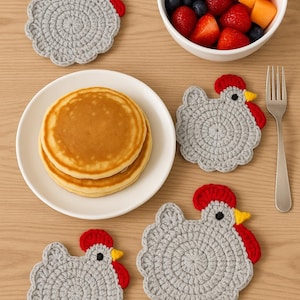May include: Four gray crochet chicken-shaped coasters with red combs and yellow beaks. A white bowl of fruit and a plate of pancakes are also on the wooden surface. A silver fork is next to the coasters.