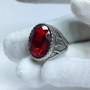 May include: A silver-toned ring featuring a large, oval-cut, faceted red gemstone set in a decorative metal band. The band displays intricate, etched geometric patterns and textured detailing along the sides, surrounding the central stone.