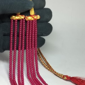 Op de afbeelding: Een close-up van een ketting met meerdere strengen kleine, rode kralen. De ketting heeft goudkleurige accenten en een rode kwast. De kralen zijn aan elkaar geregen, waardoor een waterval-effect ontstaat. De ketting wordt tegen een donkere achtergrond gehouden.