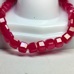 May include: A close-up of a bracelet made of faceted, deep pink beads. The beads are square-shaped with a reflective, iridescent surface. The bracelet is resting on a white surface.