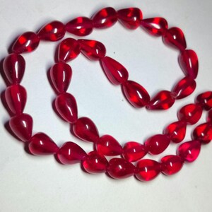 Puede incluir: Un collar de cuentas translúcidas de color rojo intenso con forma de lágrima. Las cuentas están ensartadas en un bucle continuo y presentan una superficie lisa y pulida que refleja la luz, creando un aspecto carmesí vibrante y uniforme.