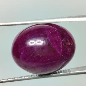 Peut inclure: Une pierre précieuse rubis de forme ovale polie, présentant une teinte rouge magenta profond et des inclusions internes visibles. La pierre est maintenue fermement entre les deux pointes métalliques d'une pince argentée, révélant sa surface lisse.