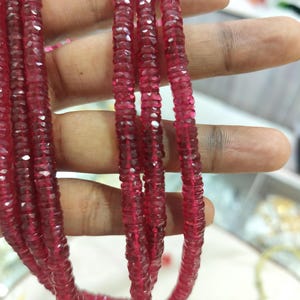 Pode incluir: Um fio de contas de pedras preciosas circulares, facetadas e de cor vermelho-profundo. As contas são pequenas, de tamanho uniforme e têm uma superfície polida e refletora que capta a luz. O fio está disposto em várias voltas.