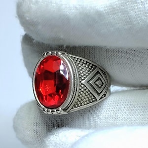 Könnte beinhalten: Ein silberfarbener Ring mit einem großen, oval geschliffenen, leuchtend roten Edelstein in der Mitte. Die Metallschiene weist an den Seiten eine texturierte, gepunktete Oberfläche auf, die durch eine geometrische Rautenprägung ergänzt wird.