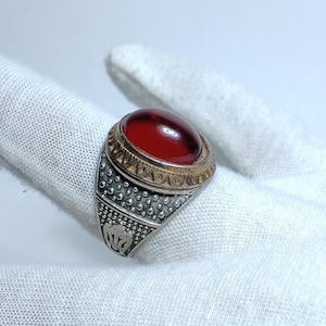 Op de afbeelding: Een zilverkleurige ring met een grote, gladde, ovaalvormige dieprode steen in een zetting. De ringband heeft een textuurrijk, korrelig oppervlak met een decoratief driehoekspatroon rond de steen en een gegraveerd embleem aan de zijkant.