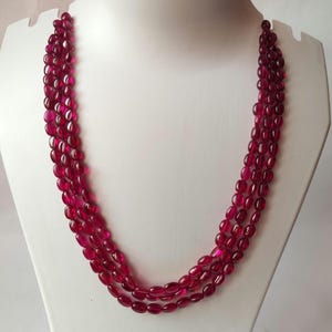 May include: A multi-strand necklace featuring three rows of small, polished, oval-shaped deep red beads. The beads are uniform in size and color, strung together to create a layered effect that drapes in a U-shape across the display form.