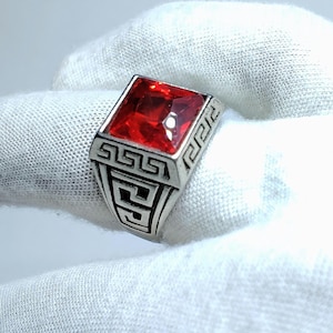 Puede incluir: Un anillo de tono plateado con una piedra preciosa roja vibrante de corte cuadrado en el centro. La banda y el engaste están decorados con un patrón geométrico de greca repetitivo grabado en la superficie metálica, dando un aspecto texturizado.
