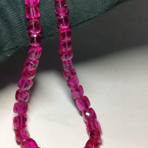 Op de afbeelding: Een ketting van gefacetteerde, felroze edelsteen kralen. De kralen zijn vierkant van vorm met meerdere facetten, wat een sprankelend effect creëert. De ketting wordt gepresenteerd tegen een donkergroene achtergrond.