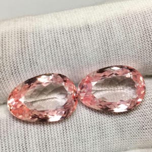 Könnte beinhalten: Zwei birnenförmige, facettierte Edelsteine in einem hellen Pfirsich-Rosa-Ton. Die Steine weisen einen Brillantschliff mit zahlreichen dreieckigen Facetten auf, die das Licht reflektieren und eine funkelnde Optik erzeugen.