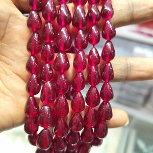 May include: A collection of deep magenta, teardrop-shaped beads strung together in multiple vertical strands. Each bead features a smooth, translucent finish that catches the light, creating a uniform and repetitive pattern of dark pink shapes.