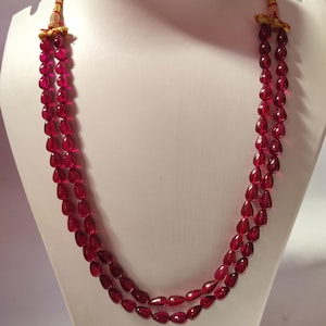Op de afbeelding: Een dubbelstrengs ketting met doorschijnende, dieprode, traanvormige kralen. De kralen zijn geregen aan een koord en aan de bovenkant vastgezet met goudkleurige metalen accenten en decoratieve draadwikkelingen bij de sluiting.