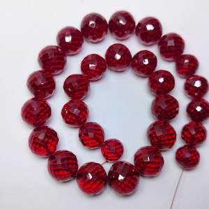 May include: A collection of round, faceted beads in a deep, translucent red color. The beads are arranged in a circular pattern, showcasing their reflective, multi-faceted surfaces that catch the light, creating a uniform and vibrant appearance.