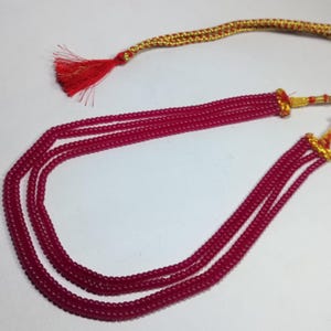 Puede incluir: Un collar de múltiples hilos con pequeñas cuentas redondas de color rojo. Tiene un cordón trenzado y detalles dorados, con una borla roja. El collar está diseñado para ser usado alrededor del cuello.