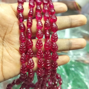 May include: A strand of translucent, deep pinkish-red gemstone beads, each carved into a detailed leaf shape with visible veins. The beads are strung together in a continuous line, showcasing a vibrant, polished finish and uniform size.